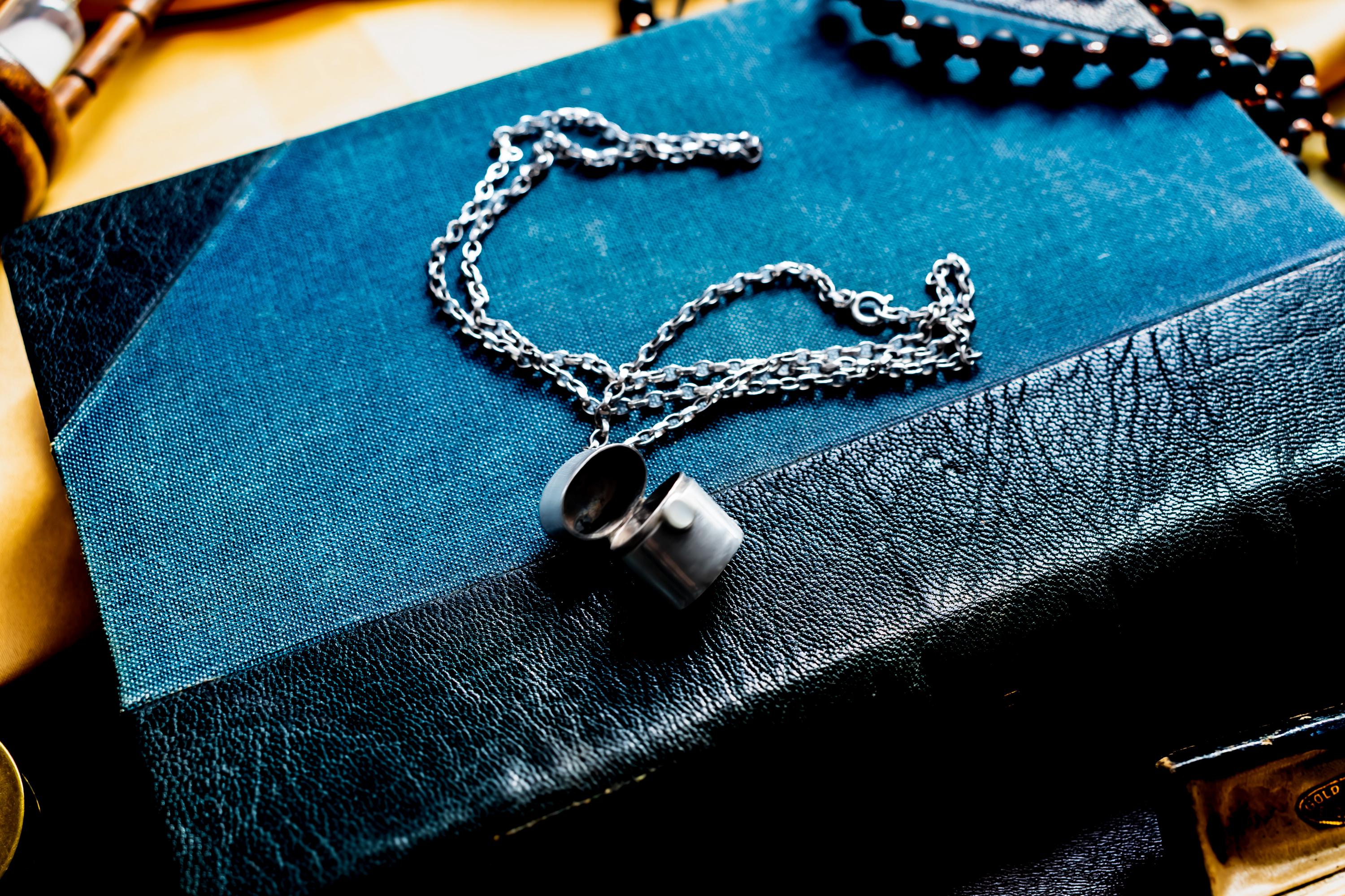 A silver-toned antique silver pill box pendant on a matching silver chain, resting on a dark blue and black textured book cover. The locket is open, revealing a hollow interior, and the chain is draped in a loose, circular pattern across the surface.