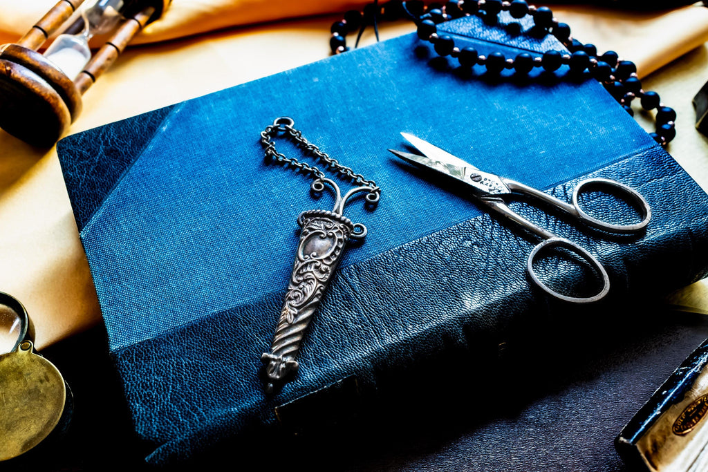 A dark blue leather-bound book featuring a silver antique metal sheath with intricate floral engravings and a matching chain, positioned next to a pair of silver-toned metal embroidery scissors with pointed blades and circular finger loops.
