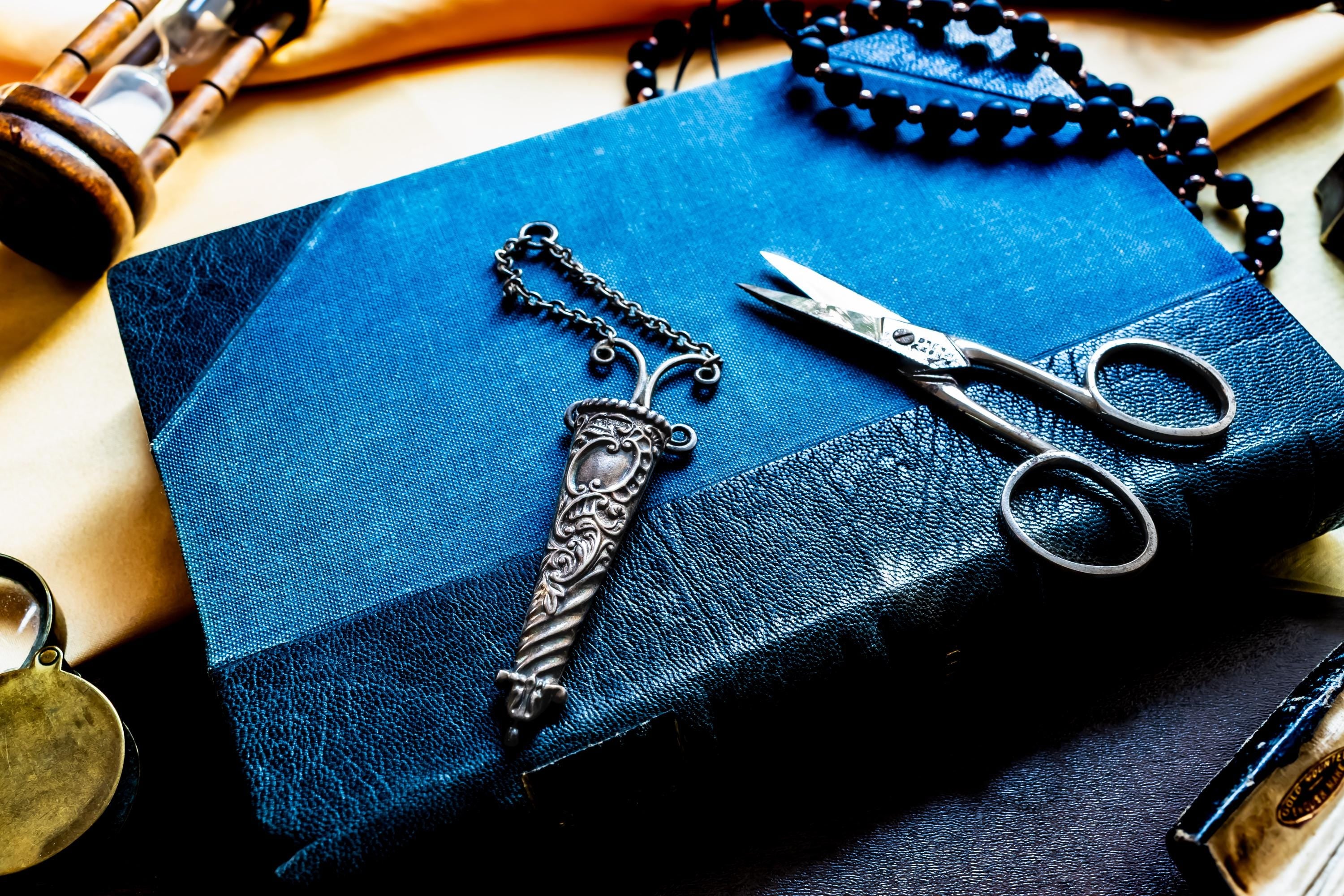 A dark blue leather-bound book featuring a silver antique metal sheath with intricate floral engravings and a matching chain, positioned next to a pair of silver-toned metal embroidery scissors with pointed blades and circular finger loops.