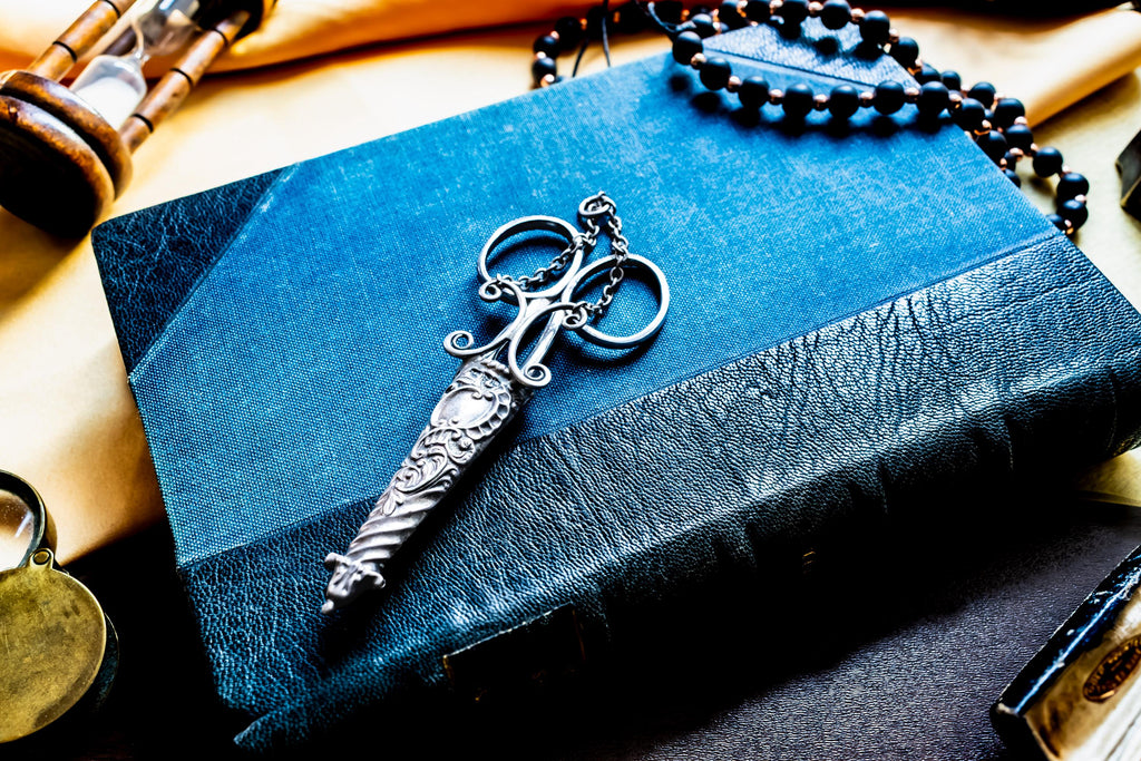 A silver antique embroidery scissors with a decorative, textured handle shaped like a dagger, resting on a rectangular blue book with a textured cover. A string of dark beads is positioned near the top of the book.