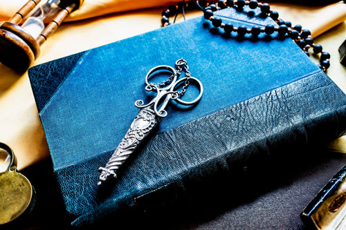 A silver antique embroidery scissors with a decorative, textured handle shaped like a dagger, resting on a rectangular blue book with a textured cover. A string of dark beads is positioned near the top of the book.