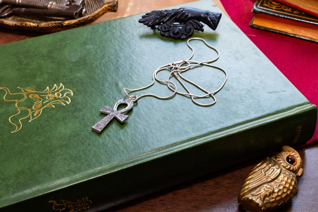 Textured sterling silver Ankh Egyptian cross pendant suspended on a fluid vintage silver snake chain. The everyday armor gothic jewelry rests on a dark green grimoire book.