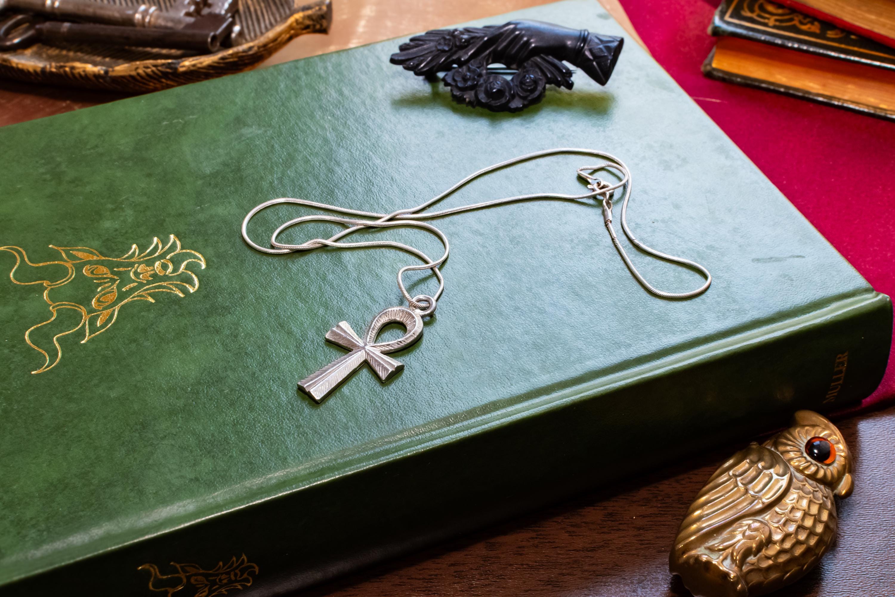 Textured sterling silver Ankh Egyptian cross pendant suspended on a fluid vintage silver snake chain. The everyday armor gothic jewelry rests on a dark green grimoire book.