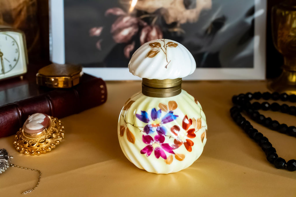 Antique pale yellow ribbed glass inkwell with hand-painted Victorian floral motifs in purple and red. The desk decor features a brass collar and a swirled white glass stopper, sitting on a writer's desk.