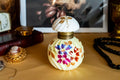 Antique pale yellow ribbed glass inkwell with hand-painted Victorian floral motifs in purple and red. The desk decor features a brass collar and a swirled white glass stopper, sitting on a writer's desk.