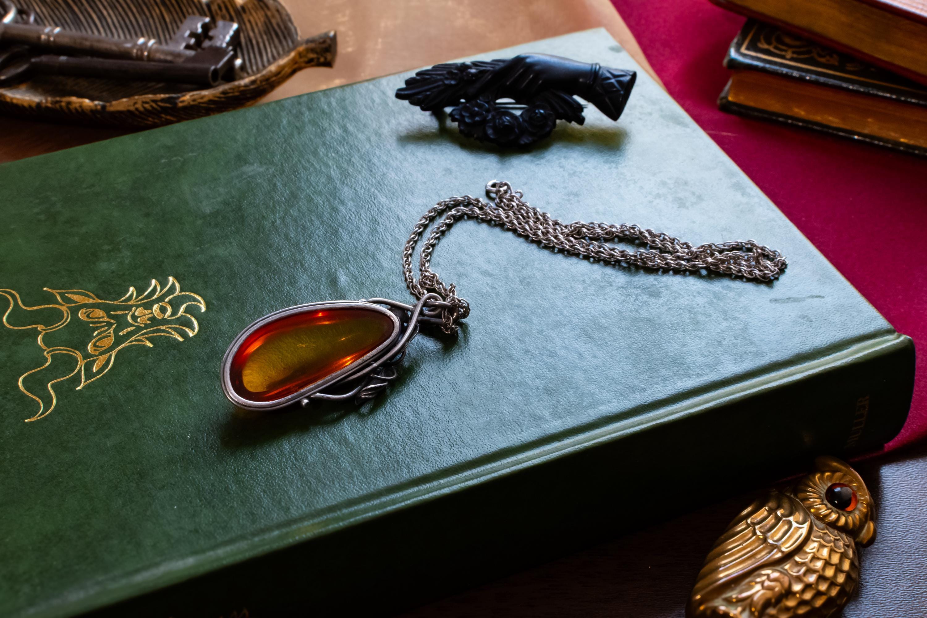Large teardrop genuine Baltic amber pendant set in an oxidized sterling silver botanical leaf design. The brown witchy gothic necklace rests on a vintage green book alongside dark academia antiquities.