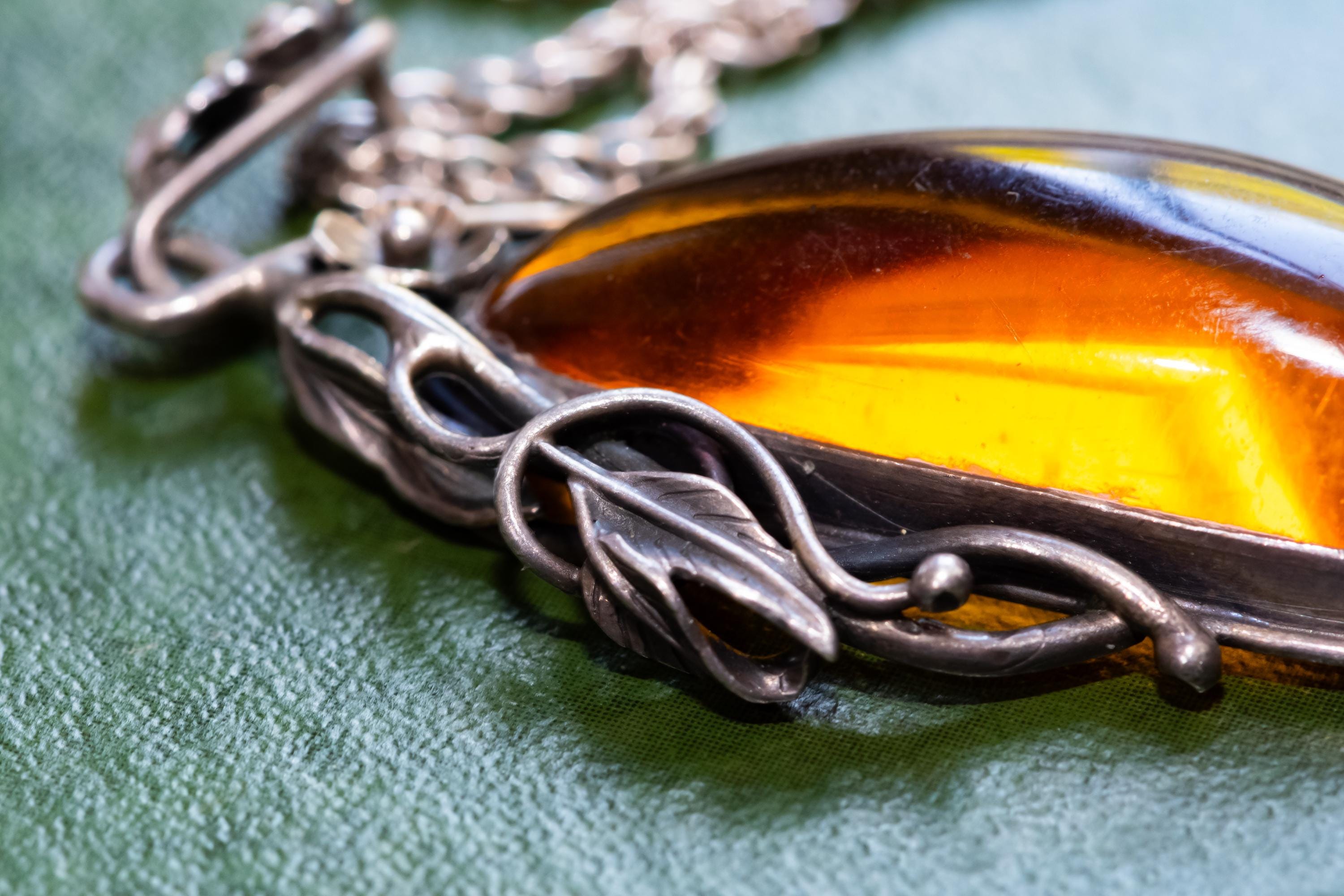 Large teardrop genuine Baltic amber pendant set in an oxidized sterling silver botanical leaf design. The brown witchy gothic necklace rests on a vintage green book alongside dark academia antiquities.