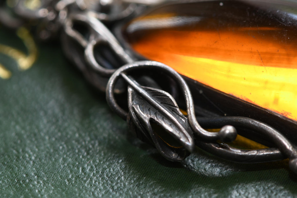 Large teardrop genuine Baltic amber pendant set in an oxidized sterling silver botanical leaf design. The brown witchy gothic necklace rests on a vintage green book alongside dark academia antiquities.