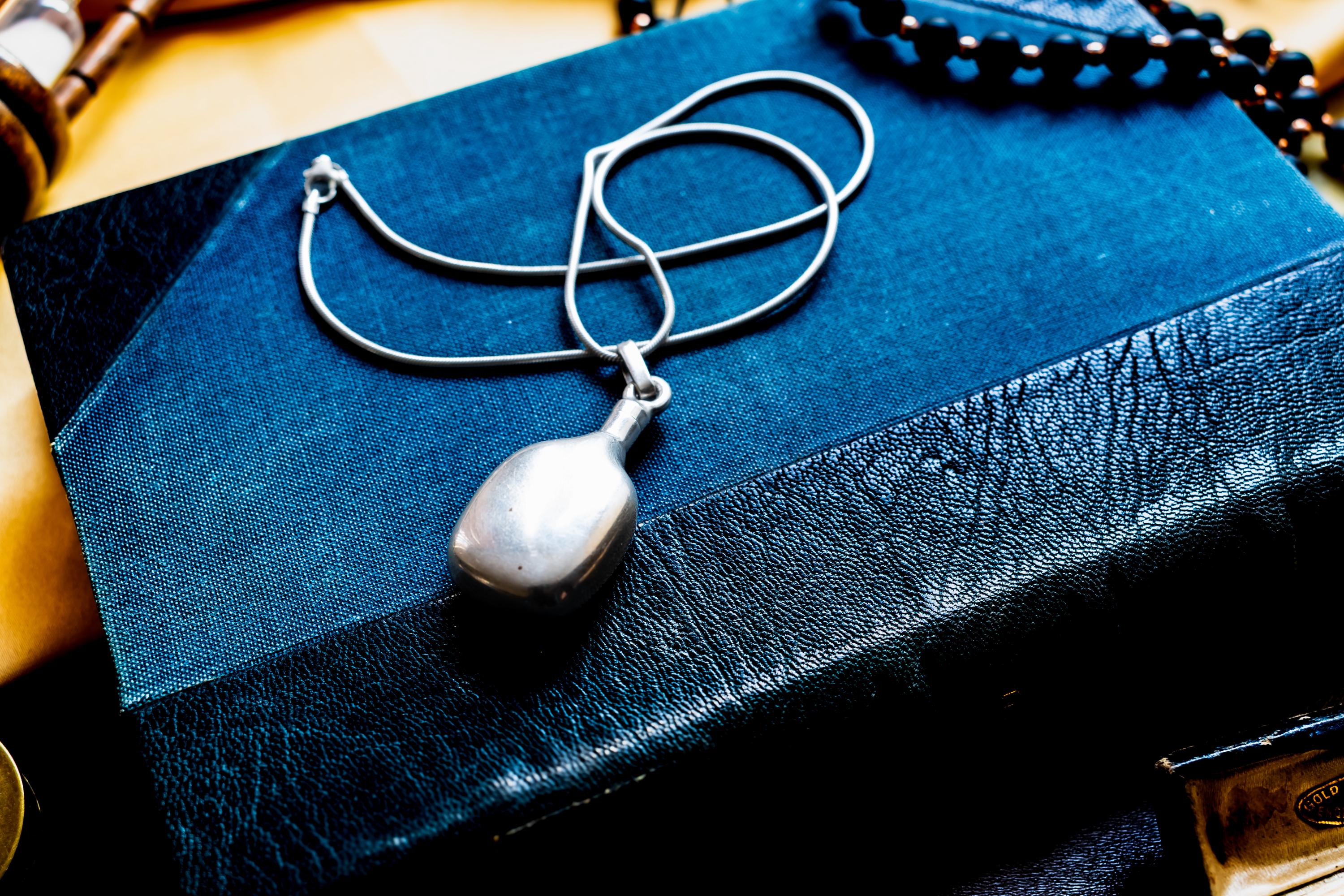 Heavy, smooth polished sterling silver antique potion flask pendant on a silver chain. The interactive Victorian scent bottle features a threaded cap and rests on a dark blue vintage book.