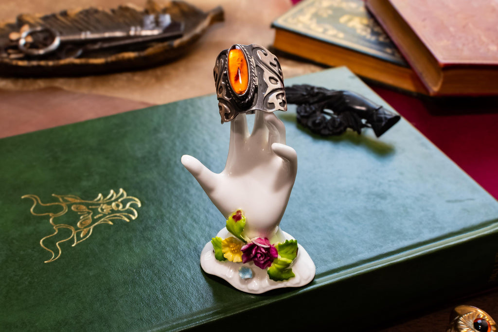 Close-up of a heavy vintage sterling silver ring featuring a dark amber cabochon top. The amber stone is hinged and slightly open to reveal a secret hidden poison locket compartment inside, set against a dark academia aesthetic background.