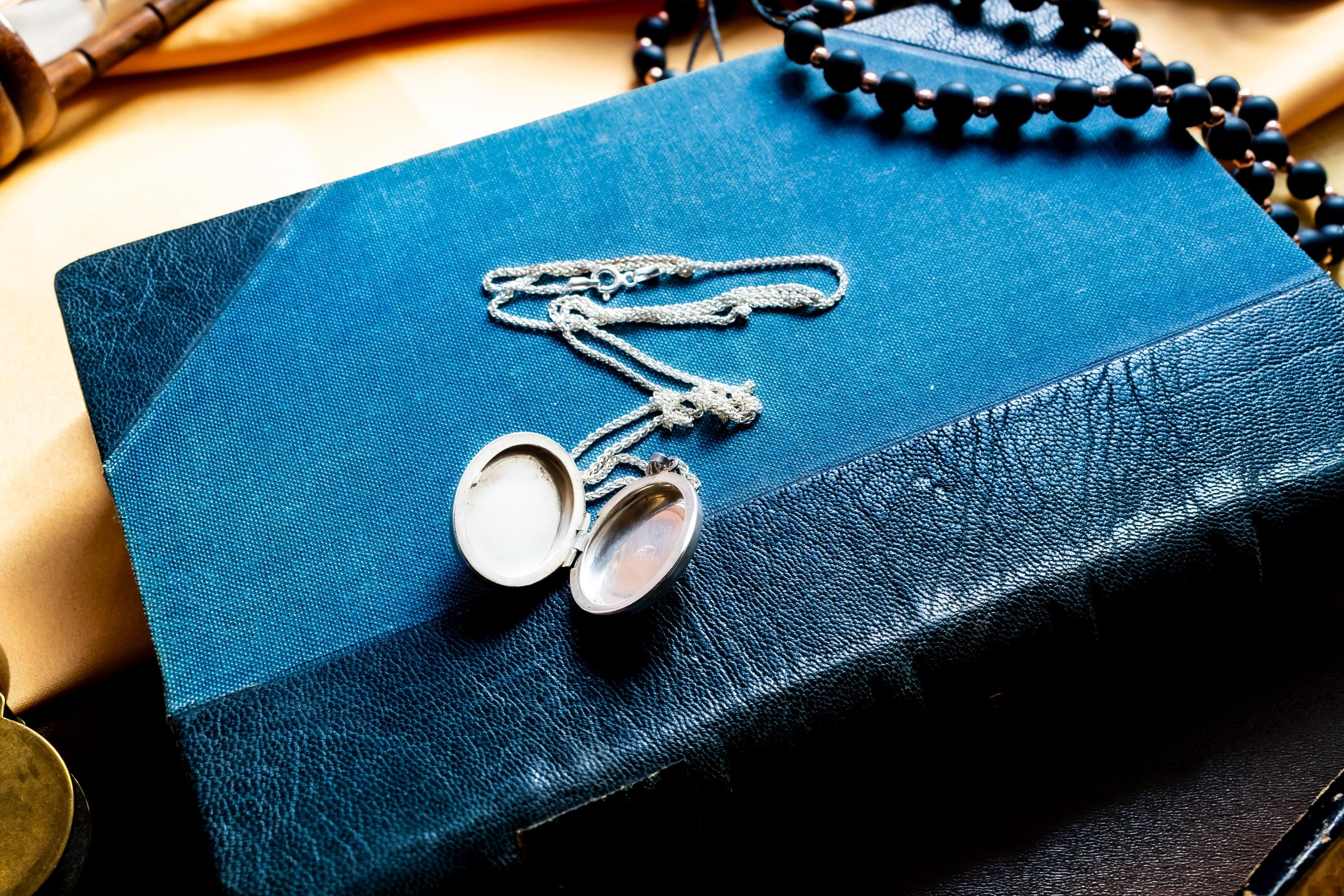 the image of an opened oval silver locket pendant featuring an intricate engraved floral pattern on its front surface. The metal has a polished, reflective finish with deep etched lines creating a swirling leaf design. A small metal loop is attached at the top.