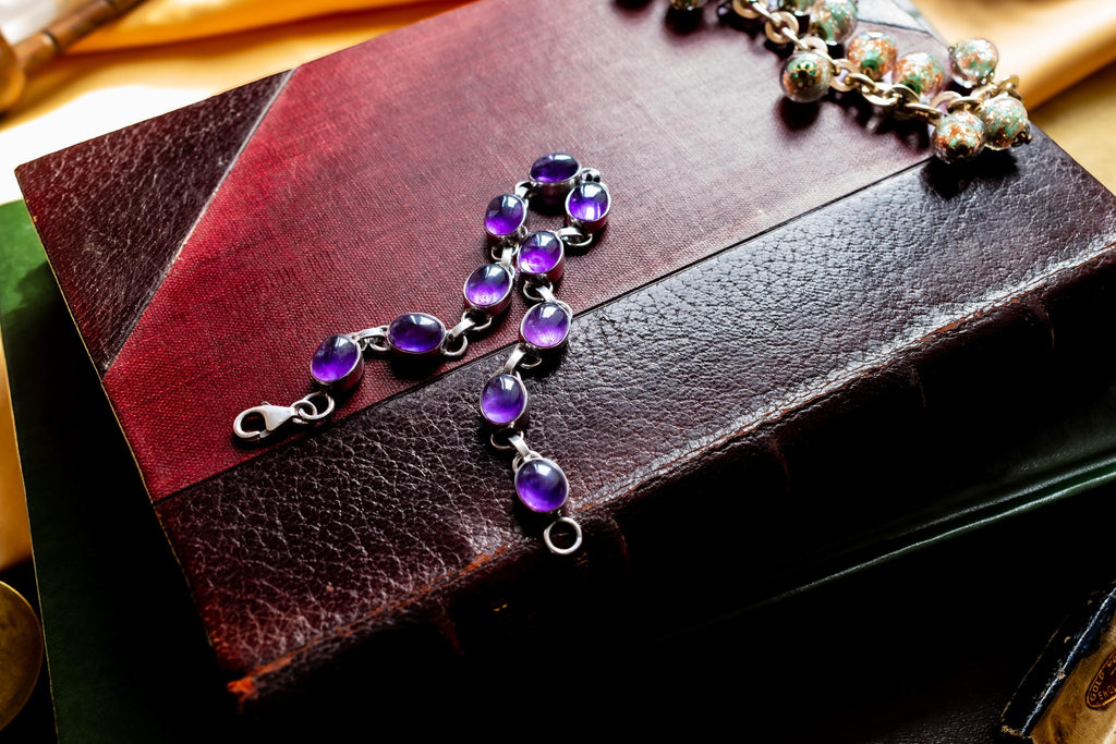 A silver bracelet featuring oval-shaped, deep purple amethyst gemstones set in polished metal links. The bracelet is draped on a red leather bound vintage book.