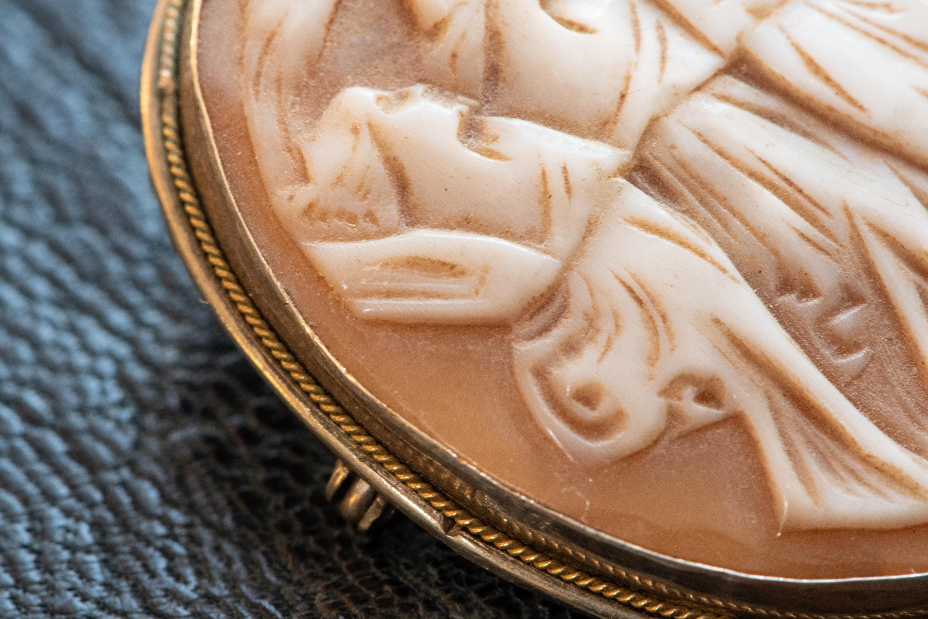 A close shot of an Antique carved shell cameo brooch depicting the Three Graces set in silver, resting on a textured blue background.