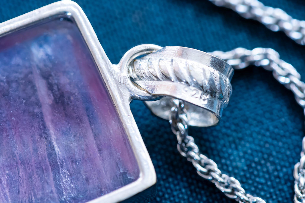 Close-up of a large, rectangular Kunzite gemstone pendant with natural pink and violet banding, set in 925 sterling silver and resting on a textured blue book.
