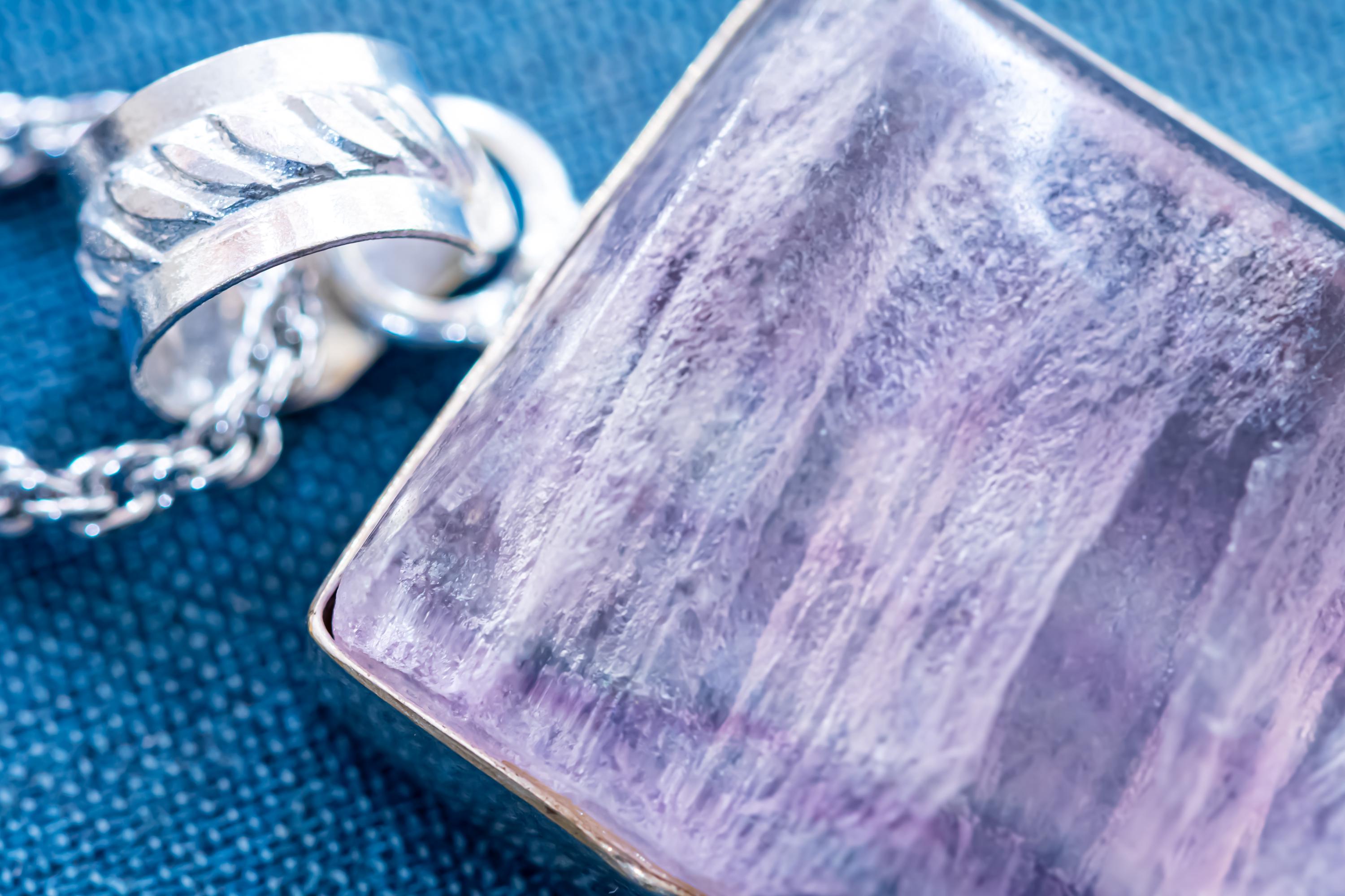 Close-up of a large, rectangular Kunzite gemstone pendant with natural pink and violet banding, set in 925 sterling silver and resting on a textured blue book.