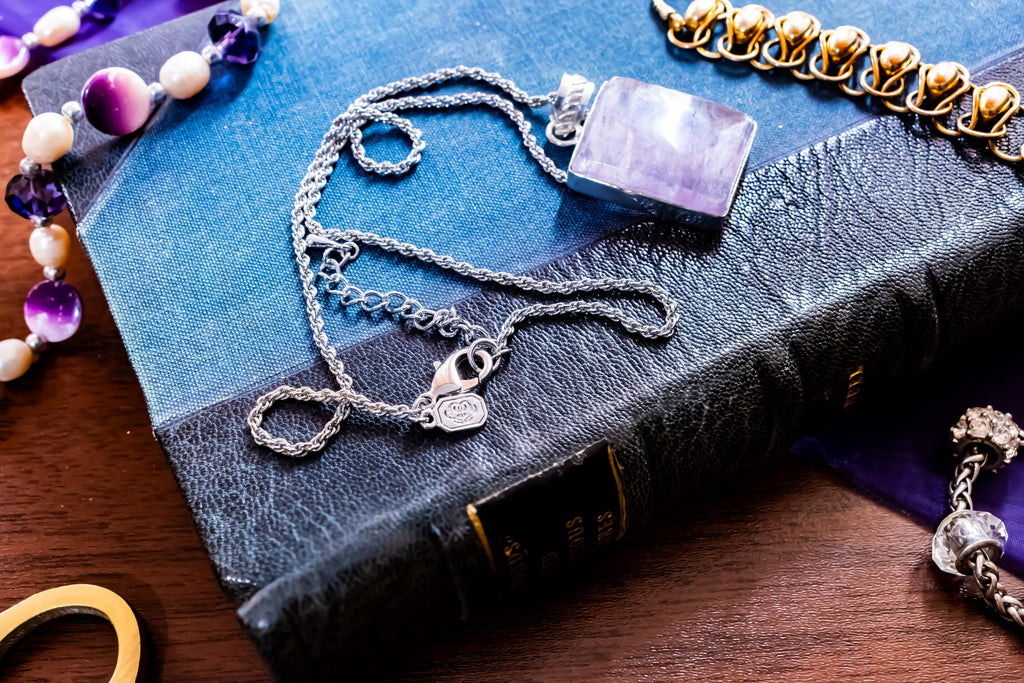 Close-up of a large, rectangular Kunzite gemstone pendant with natural pink and violet banding, set in 925 sterling silver and resting on a textured blue book.