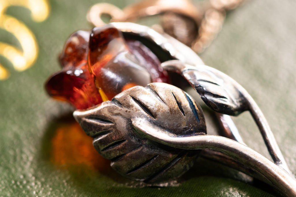 A vintage 925 sterling silver necklace featuring a carved Baltic amber rose pendant, beautifully staged resting on the cover of an antique green book with a dark carved brooch in the background.