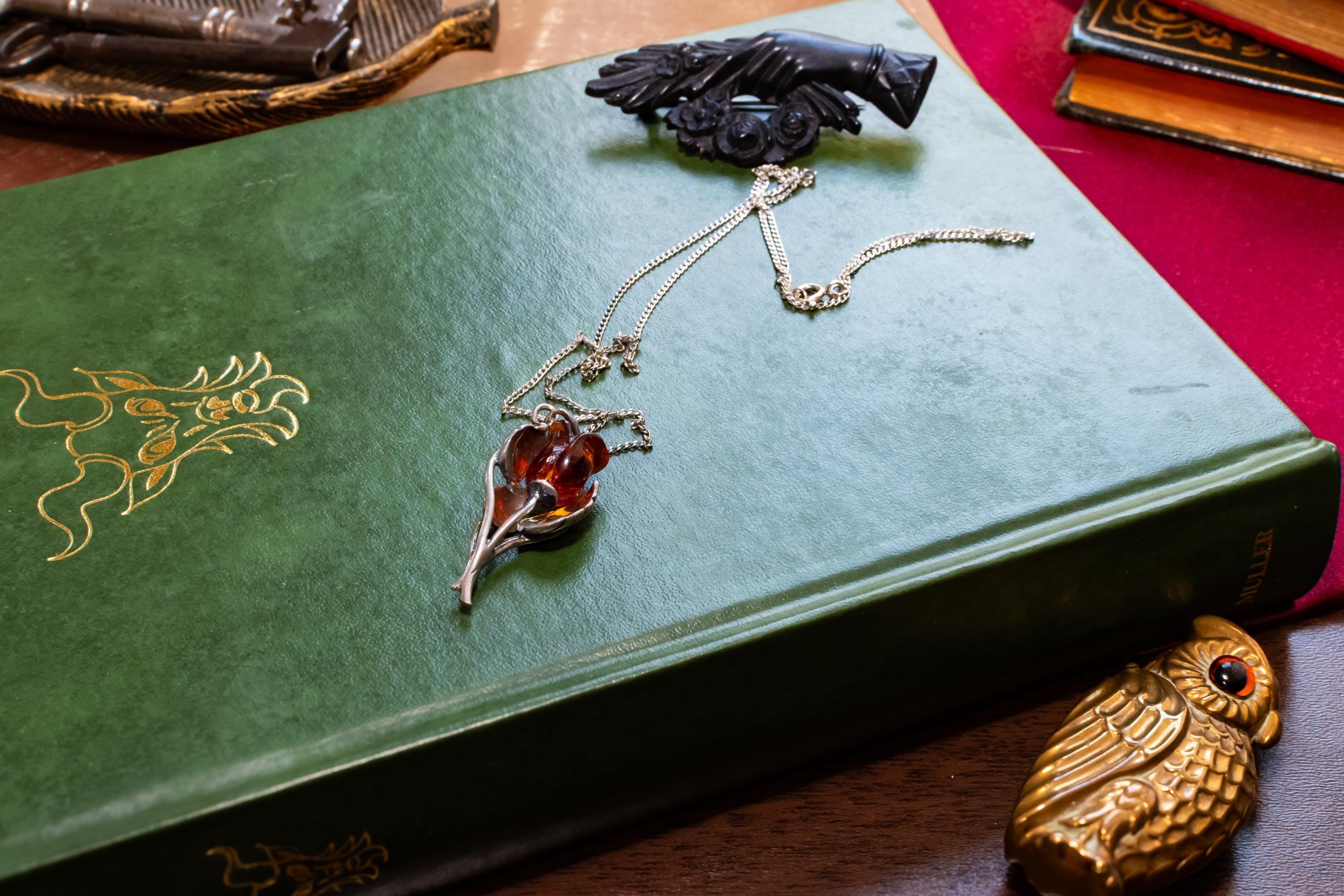 A vintage 925 sterling silver necklace featuring a carved Baltic amber rose pendant, beautifully staged resting on the cover of an antique green book with a dark carved brooch in the background.