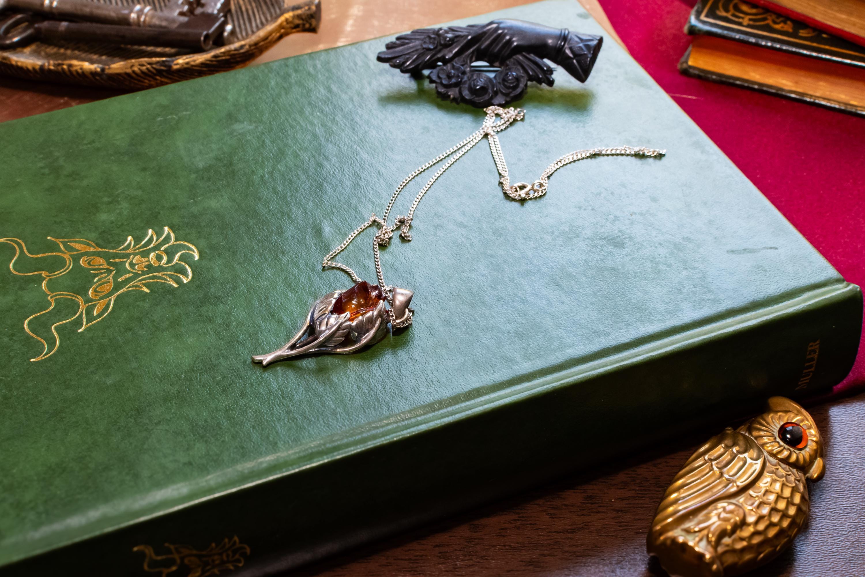 A vintage 925 sterling silver necklace featuring a carved Baltic amber rose pendant, beautifully staged resting on the cover of an antique green book with a dark carved brooch in the background.