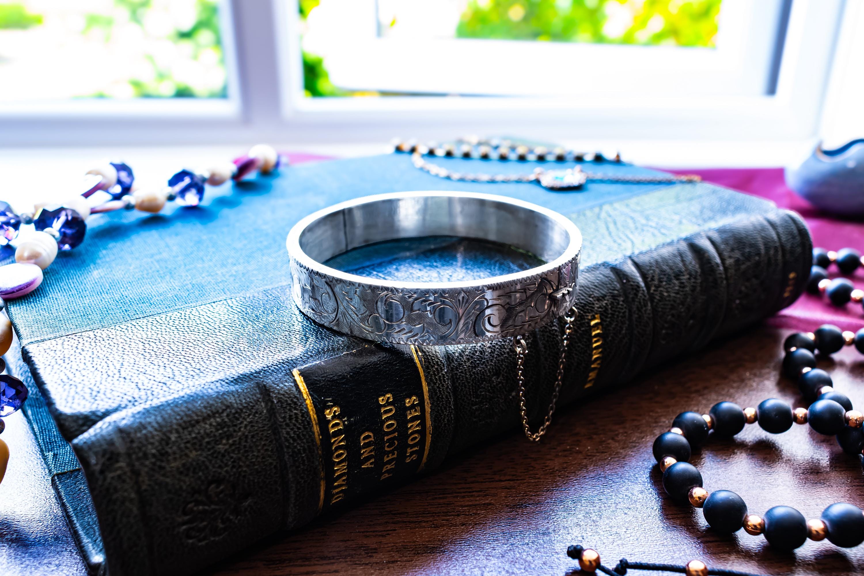 A vintage 1979 Sheffield sterling silver hinged cuff bracelet featuring intricate scrollwork engraving and a delicate safety chain, resting on the spine of an antique dark green book titled 'Diamonds and Precious Stones'.