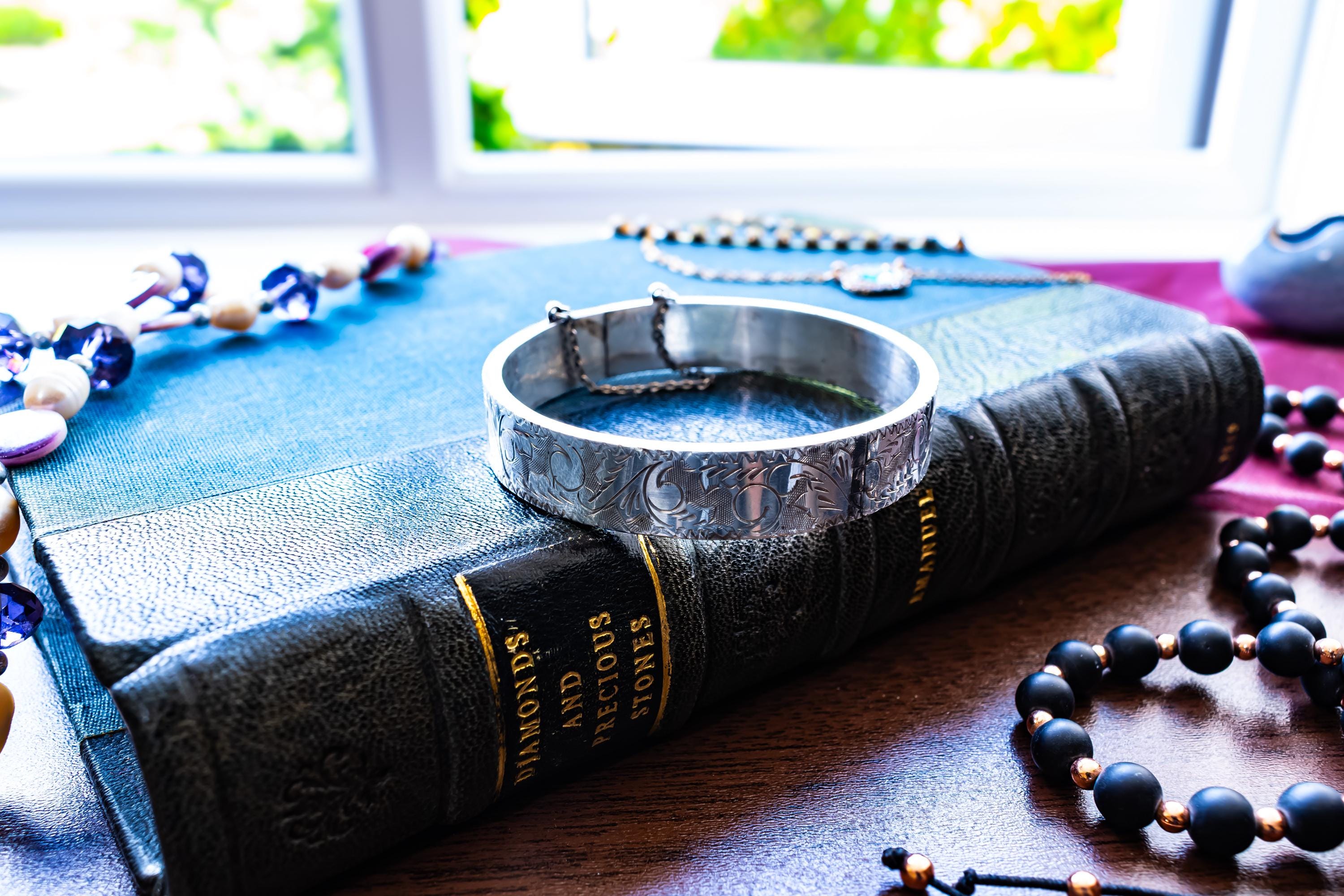 A vintage 1979 Sheffield sterling silver hinged cuff bracelet featuring intricate scrollwork engraving and a delicate safety chain, resting on the spine of an antique dark green book titled 'Diamonds and Precious Stones'.