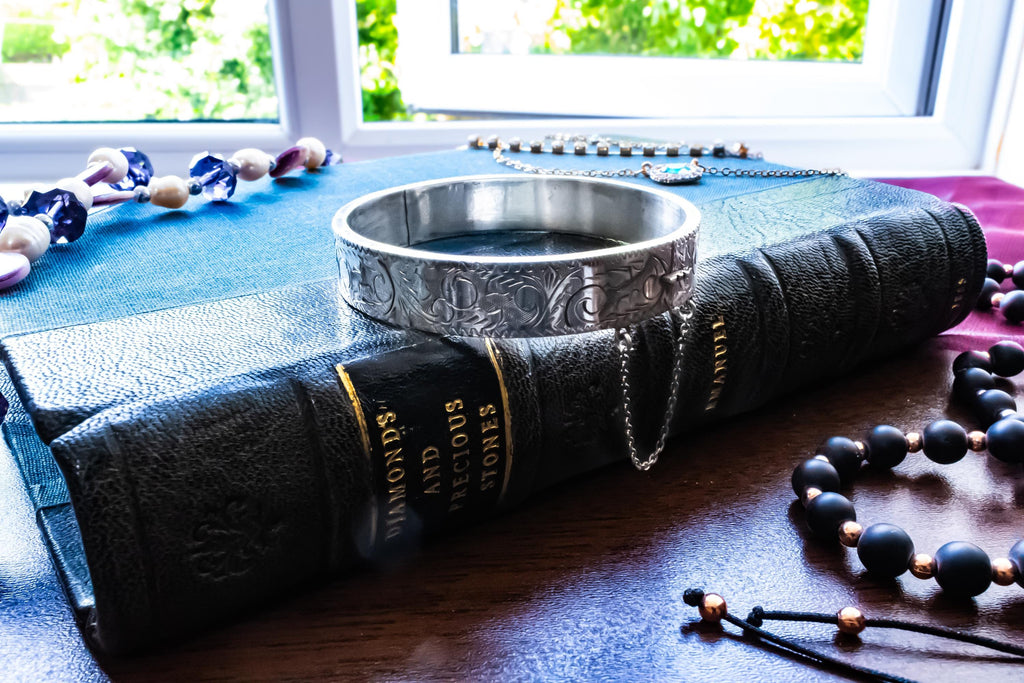 A vintage 1979 Sheffield sterling silver hinged cuff bracelet featuring intricate scrollwork engraving and a delicate safety chain, resting on the spine of an antique dark green book titled 'Diamonds and Precious Stones'.
