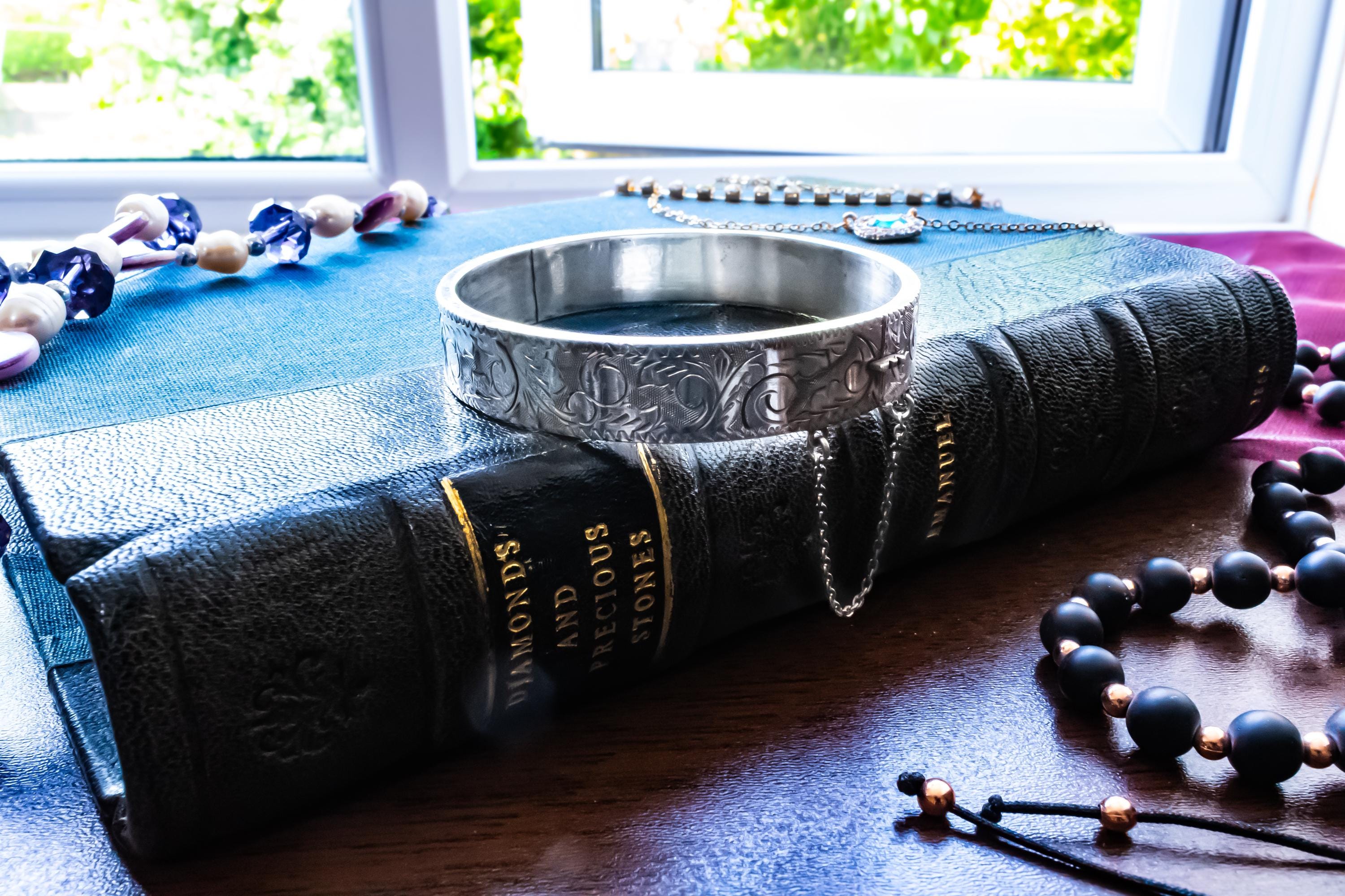 A vintage 1979 Sheffield sterling silver hinged cuff bracelet featuring intricate scrollwork engraving and a delicate safety chain, resting on the spine of an antique dark green book titled 'Diamonds and Precious Stones'.