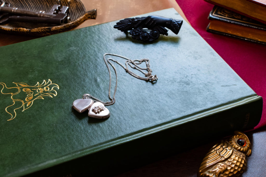 A vintage silver heart locket necklace featuring a small embossed teddy bear design, resting beautifully on a green antique book with gold embossing. A small brass owl and a carved black hand brooch sit in the background.