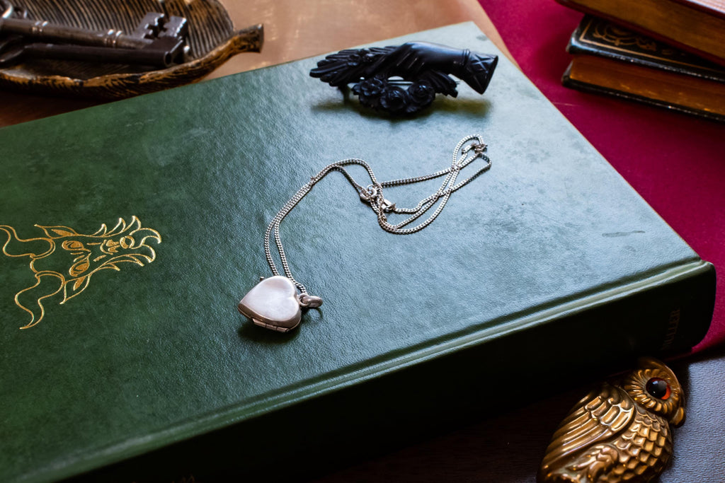 A vintage silver heart locket necklace featuring a small embossed teddy bear design, resting beautifully on a green antique book with gold embossing. A small brass owl and a carved black hand brooch sit in the background.
