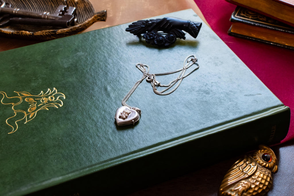 A vintage silver heart locket necklace featuring a small embossed teddy bear design, resting beautifully on a green antique book with gold embossing. A small brass owl and a carved black hand brooch sit in the background.