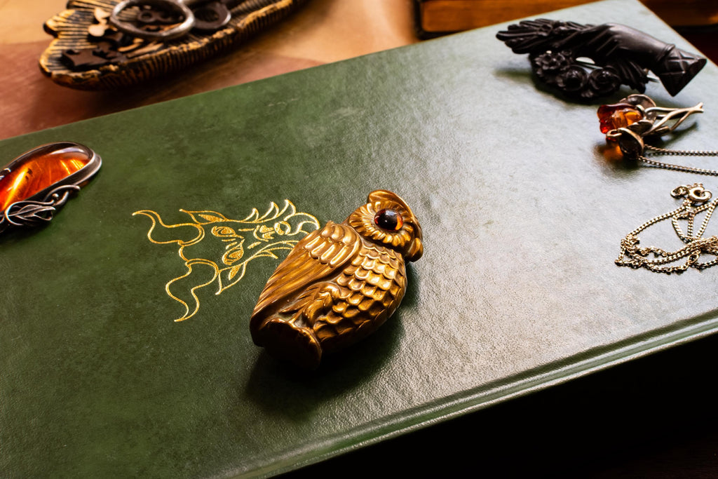 A heavy brass vesta case shaped like an owl with glowing amber glass eyes, resting on the cover of a vintage green book with gold embossing. Assorted vintage jewelry sits softly out of focus in the background.