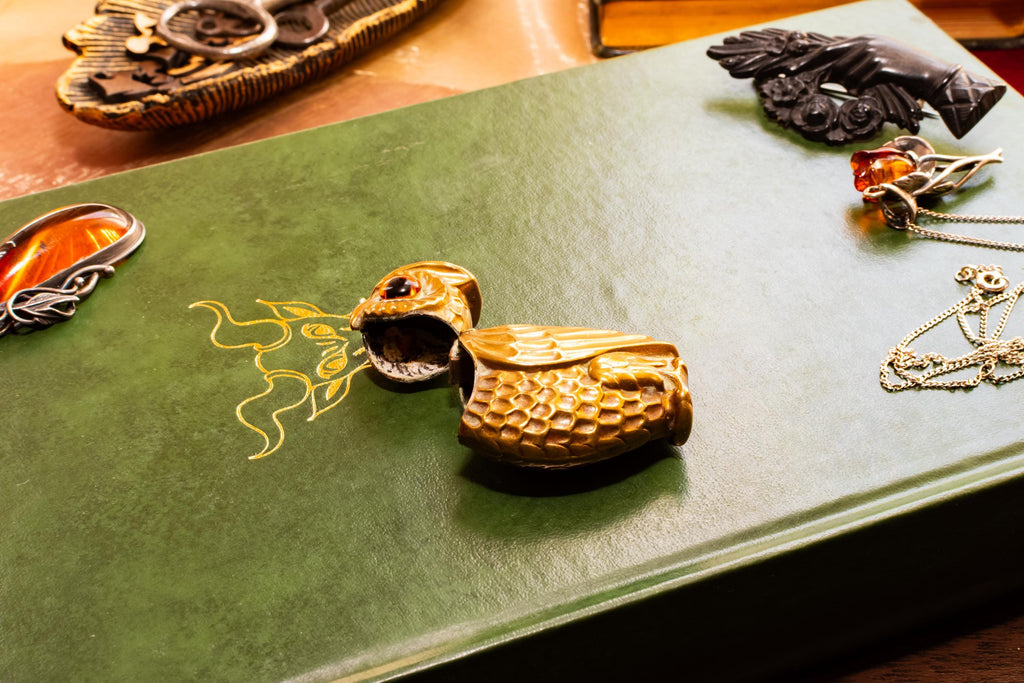 A heavy brass vesta case shaped like an owl with glowing amber glass eyes, resting on the cover of a vintage green book with gold embossing. Assorted vintage jewelry sits softly out of focus in the background.