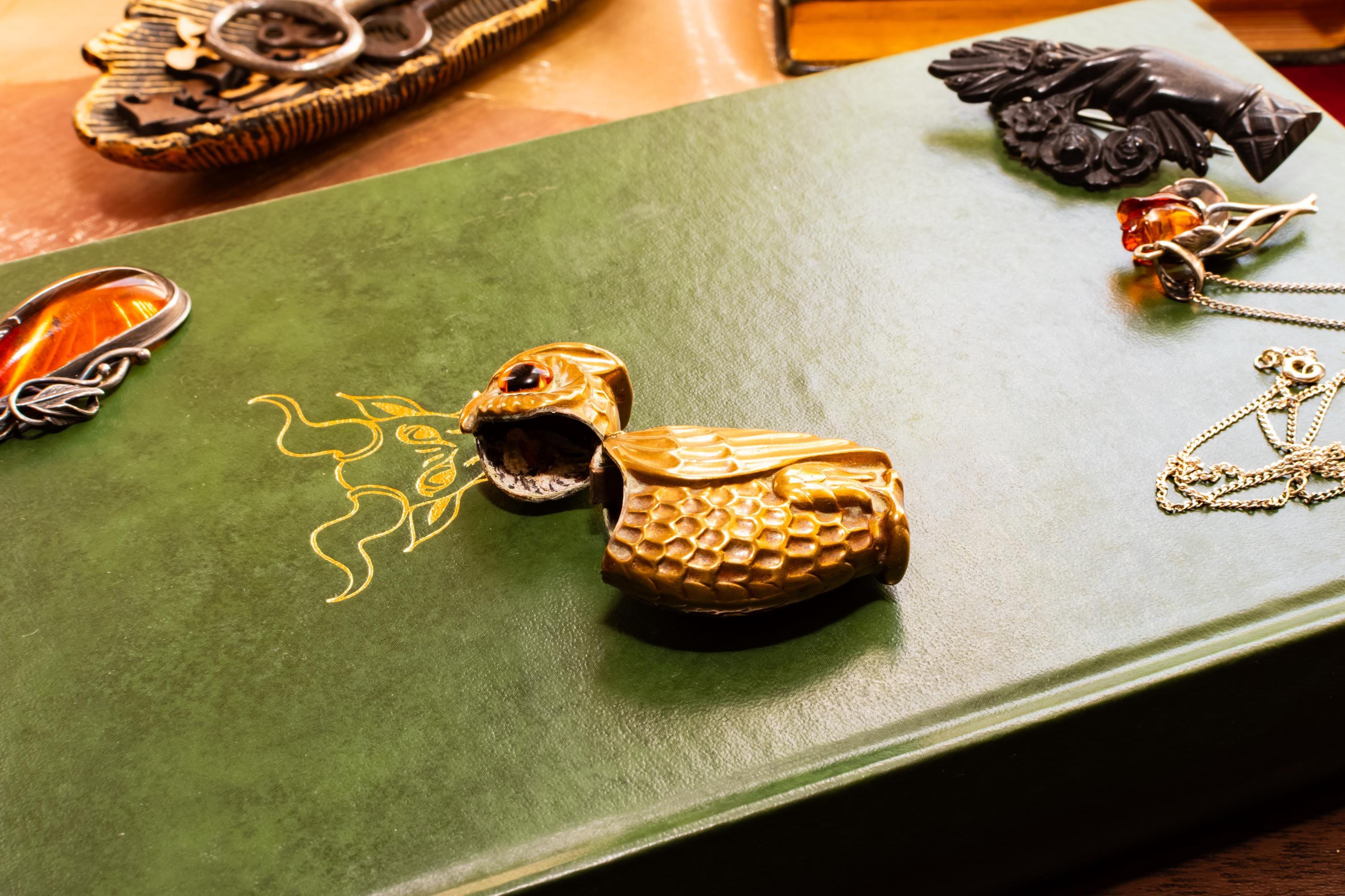 A heavy brass vesta case shaped like an owl with glowing amber glass eyes, resting on the cover of a vintage green book with gold embossing. Assorted vintage jewelry sits softly out of focus in the background.