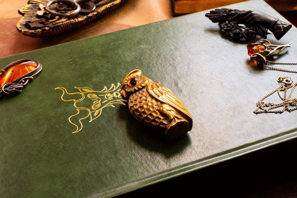 A heavy brass vesta case shaped like an owl with glowing amber glass eyes, resting on the cover of a vintage green book with gold embossing. Assorted vintage jewelry sits softly out of focus in the background.