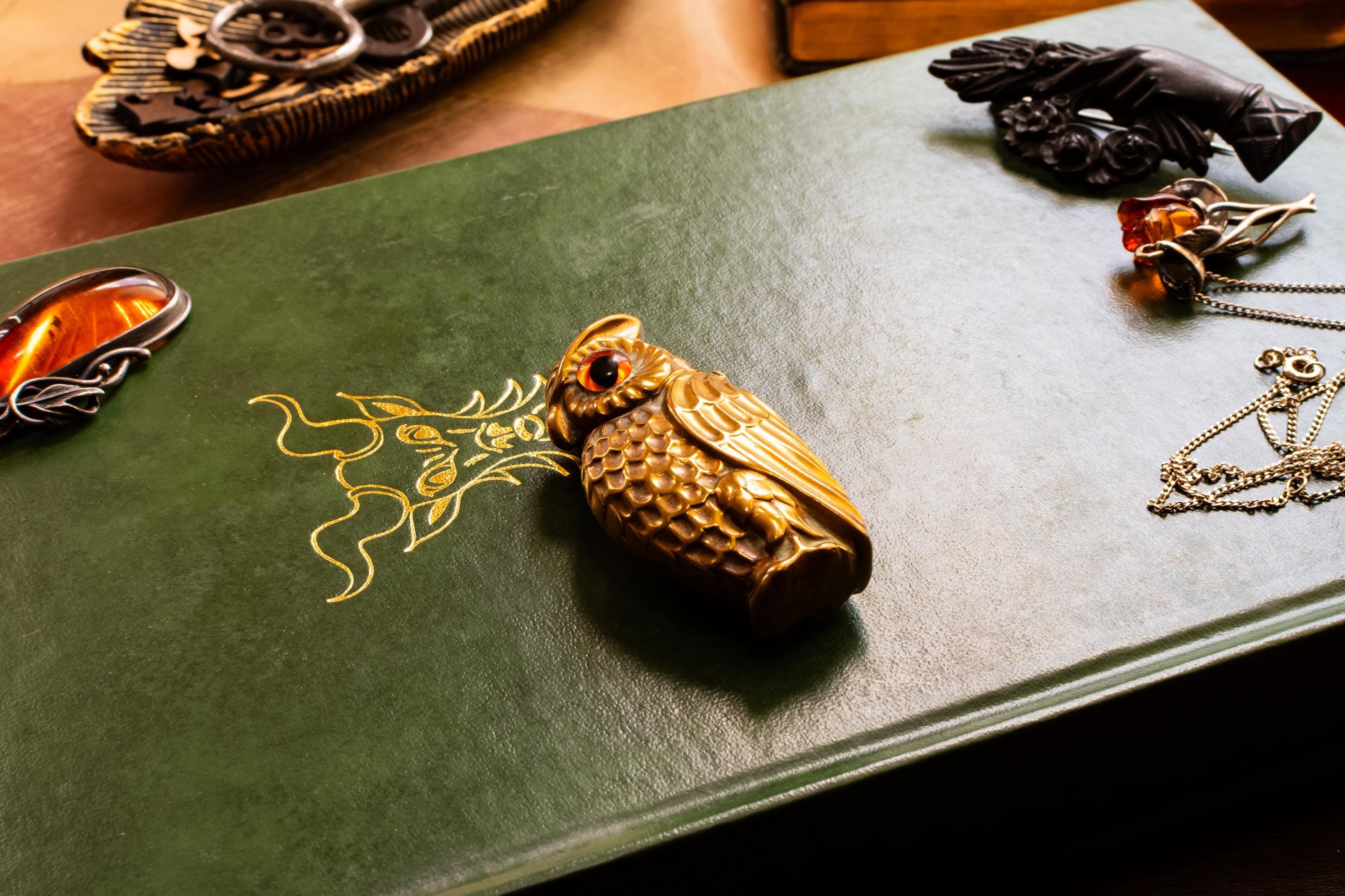 A heavy brass vesta case shaped like an owl with glowing amber glass eyes, resting on the cover of a vintage green book with gold embossing. Assorted vintage jewelry sits softly out of focus in the background.