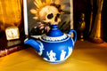 A Matte blue and white team pot resting on a gold material. The teapot has white neoclassical raised images around the pot.