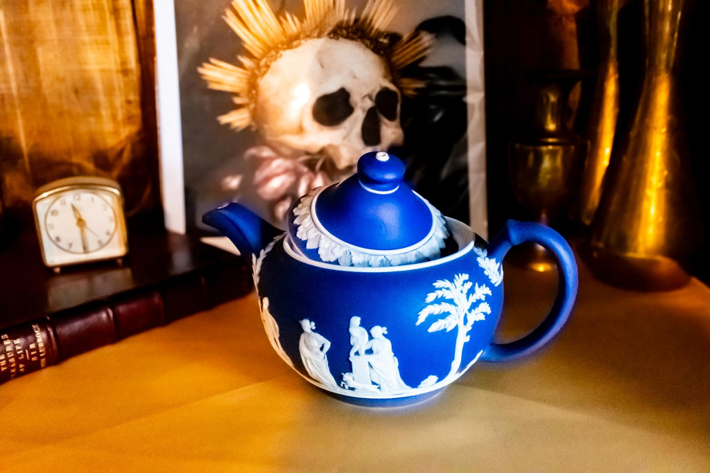 A Matte blue and white team pot resting on a gold material. The teapot has white neoclassical raised images around the pot.