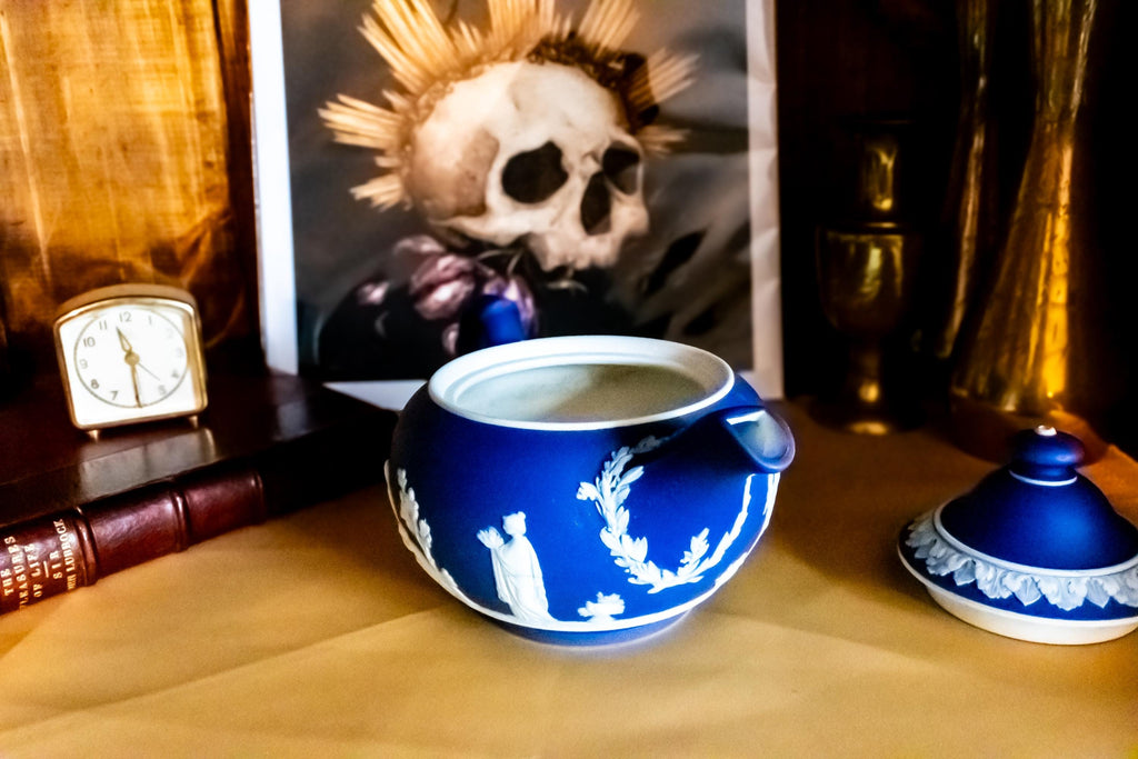 A Matte blue and white team pot resting on a gold material. The teapot has white neoclassical raised images around the pot.