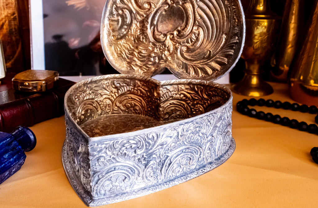 A solid sterling silver hand engraved jewelry box open to show the gold coloring of the inside. The shape of the jewelry box is a hard. It has engravings on the top and on the sides.