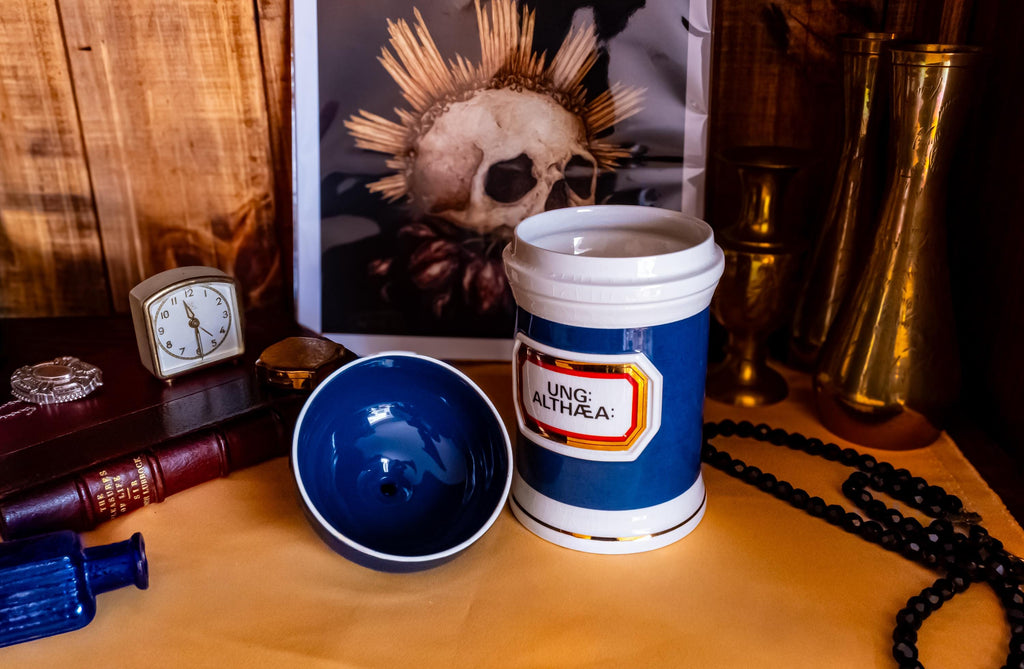 Antique apothecary blue and white jar with a label that says Ung: Althaea. The label is surrounded by gold foil. The white jar has a blue porcelain lid on its side.
