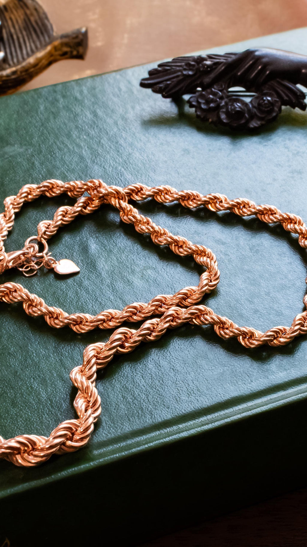 A thick, vintage rose gold vermeil rope chain necklace, resting beautifully on a green antique book with gold dragon embossing. Assorted vintage jewelry sits softly out of focus in the background.