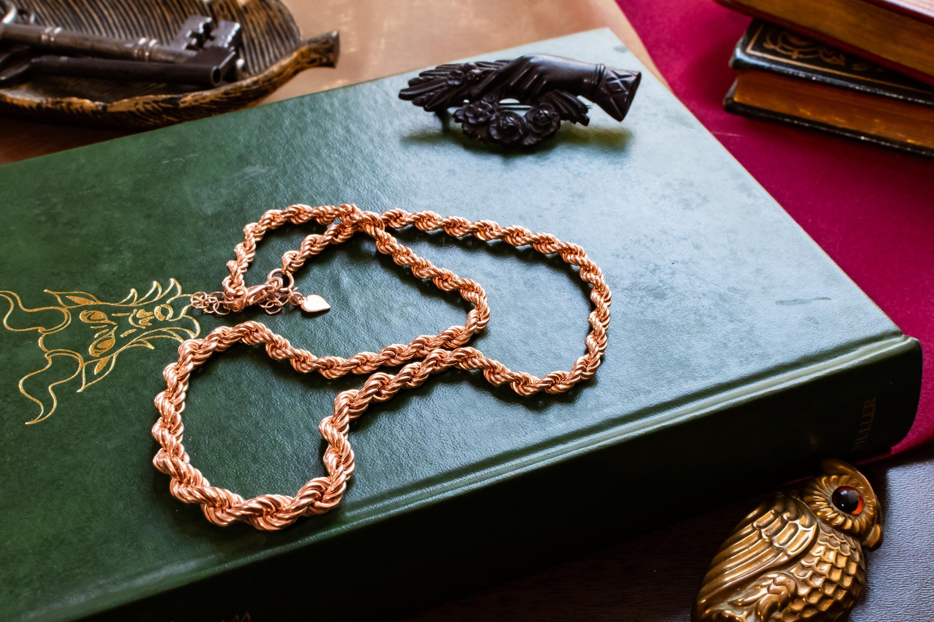 A thick, vintage rose gold vermeil rope chain necklace, resting beautifully on a green antique book with gold dragon embossing. Assorted vintage jewelry sits softly out of focus in the background.