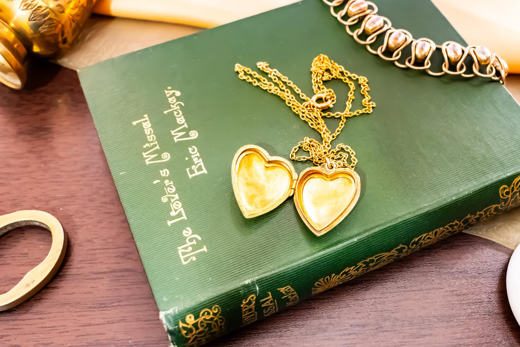An antique 9ct gold back and front heart-shaped locket featuring a delicate hand-engraved swallow, resting romantically on a green vintage book titled "The Lover's Missal".