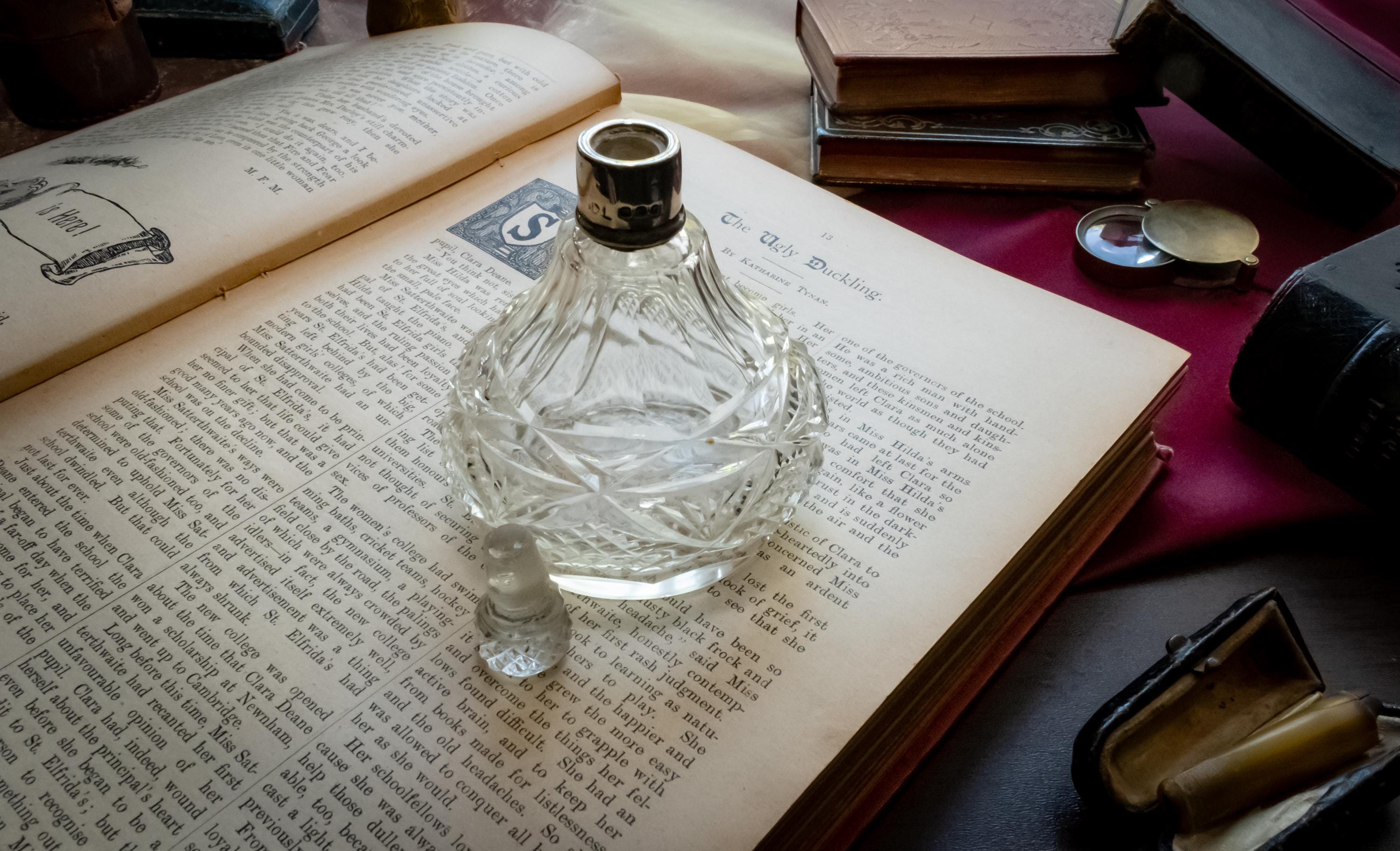 An antique heavy cut glass scent bottle with a sterling silver collar and crystal stopper, sitting on an open vintage book surrounded by dark academia props