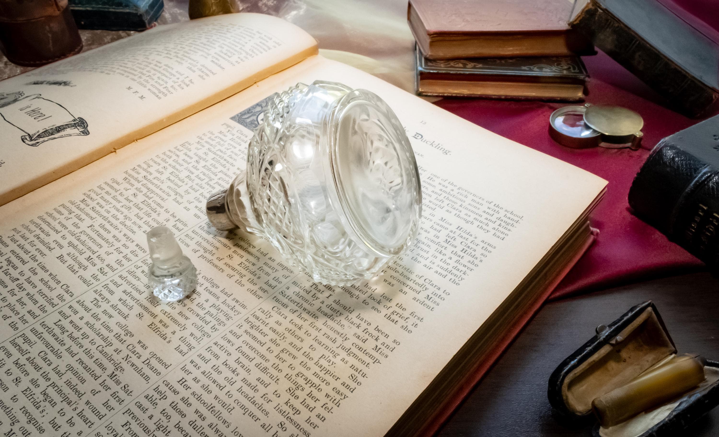 An antique heavy cut glass scent bottle with a sterling silver collar and crystal stopper, sitting on an open vintage book surrounded by dark academia props