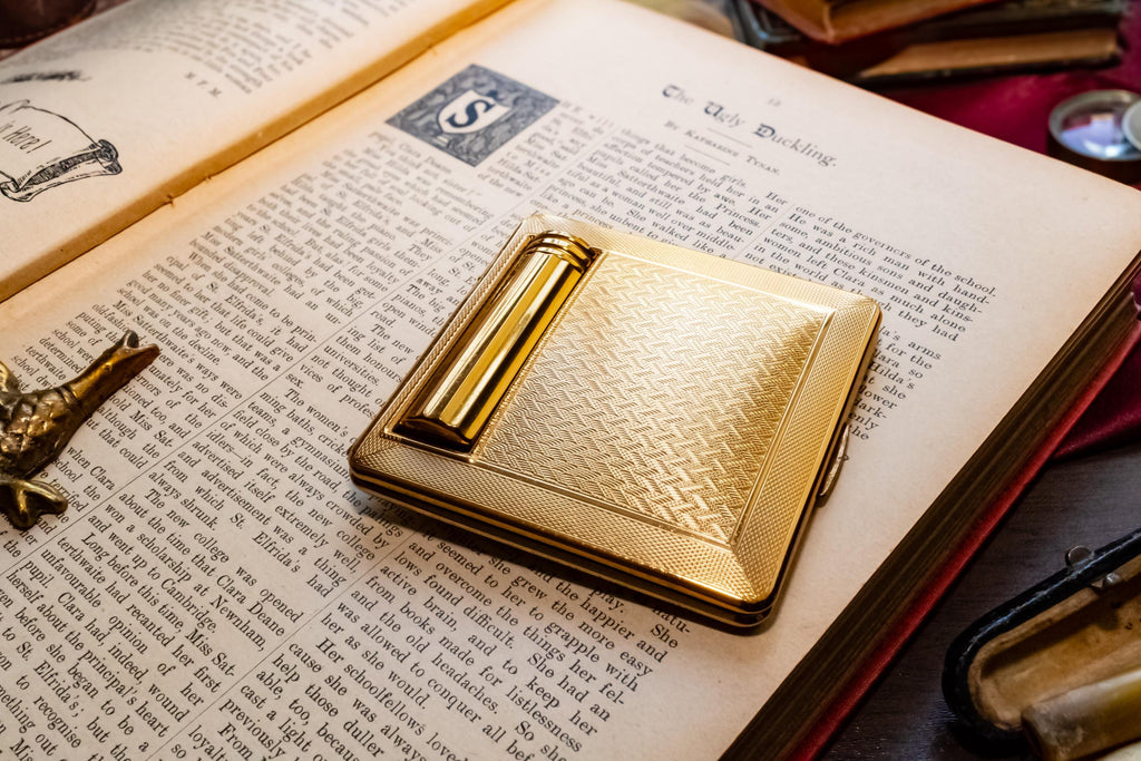 A gleaming gold vintage Kigu of London rectangular compact with a basketweave pattern and integrated lipstick holder, resting on an open antique book.