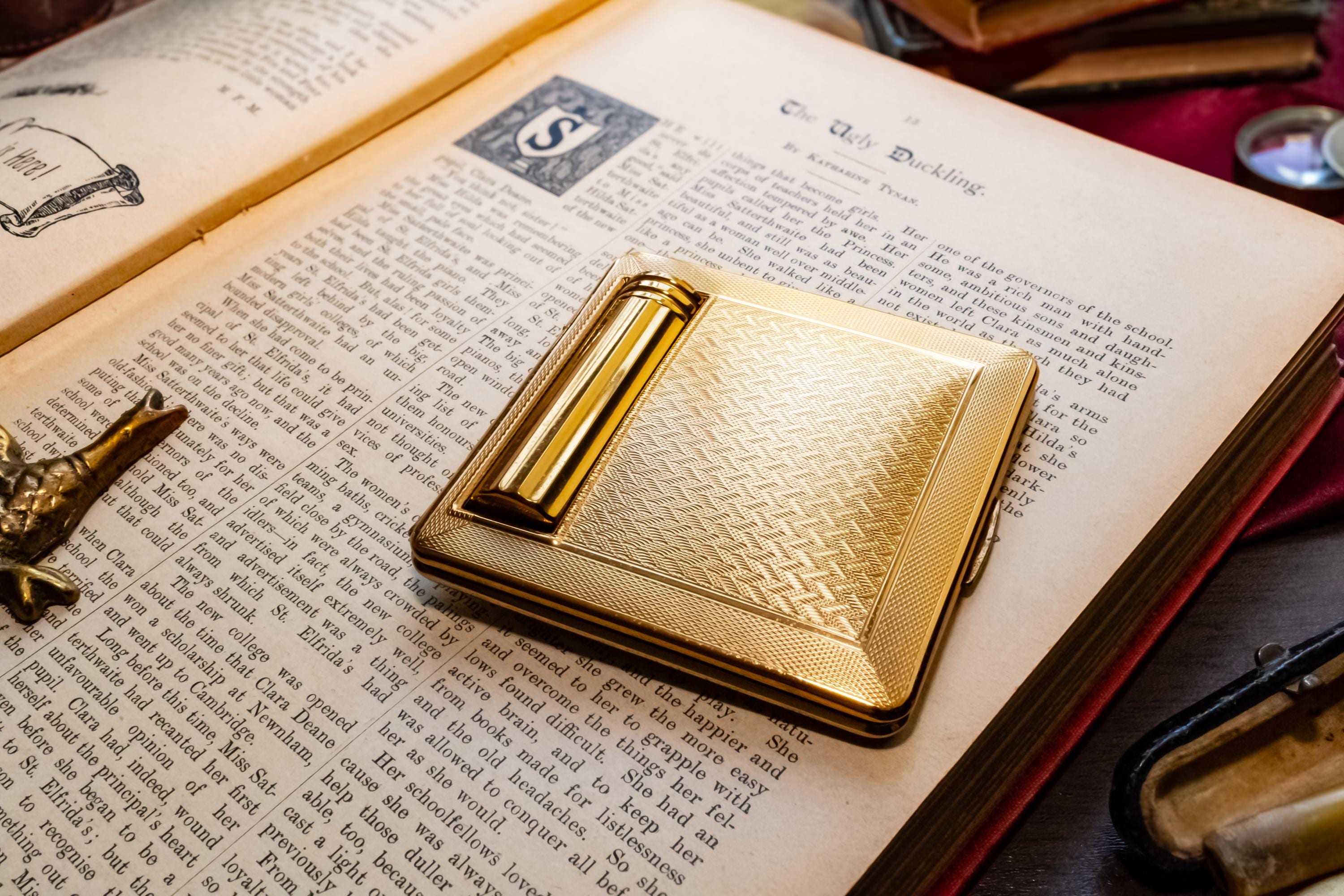 A gleaming gold vintage Kigu of London rectangular compact with a basketweave pattern and integrated lipstick holder, resting on an open antique book.
