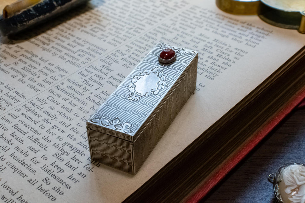 A rectangular silver-toned metal box with engraved floral detailing and a small red gemstone on top, resting on the pages of an open antique book.