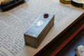 A rectangular silver-toned metal box with engraved floral detailing and a small red gemstone on top, resting on the pages of an open antique book.