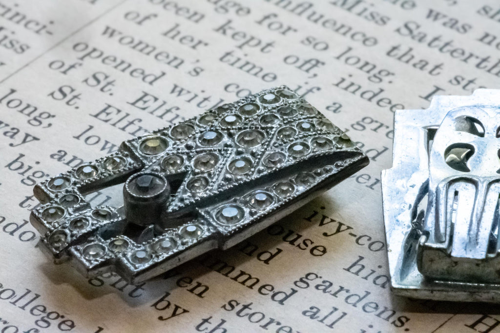 A silver-toned vintage brooch with intricate diamond-cut detailing and a central screw-like accent, displayed on an open book page next to its back or reverse side.